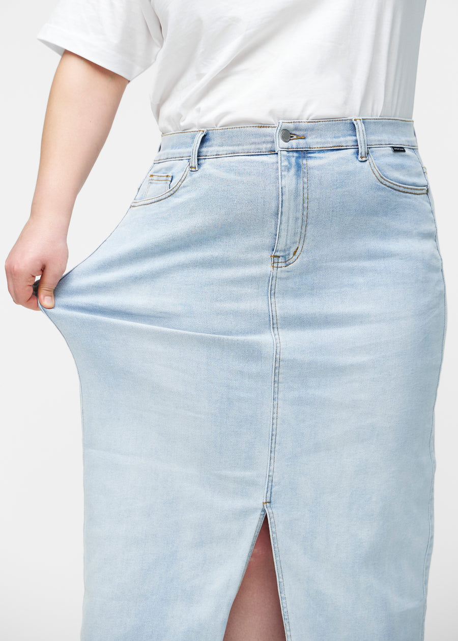 Plus-size model strækker den lange, lyseblå denimnederdel i farven Waves ved hoften for at vise materialets stretch.