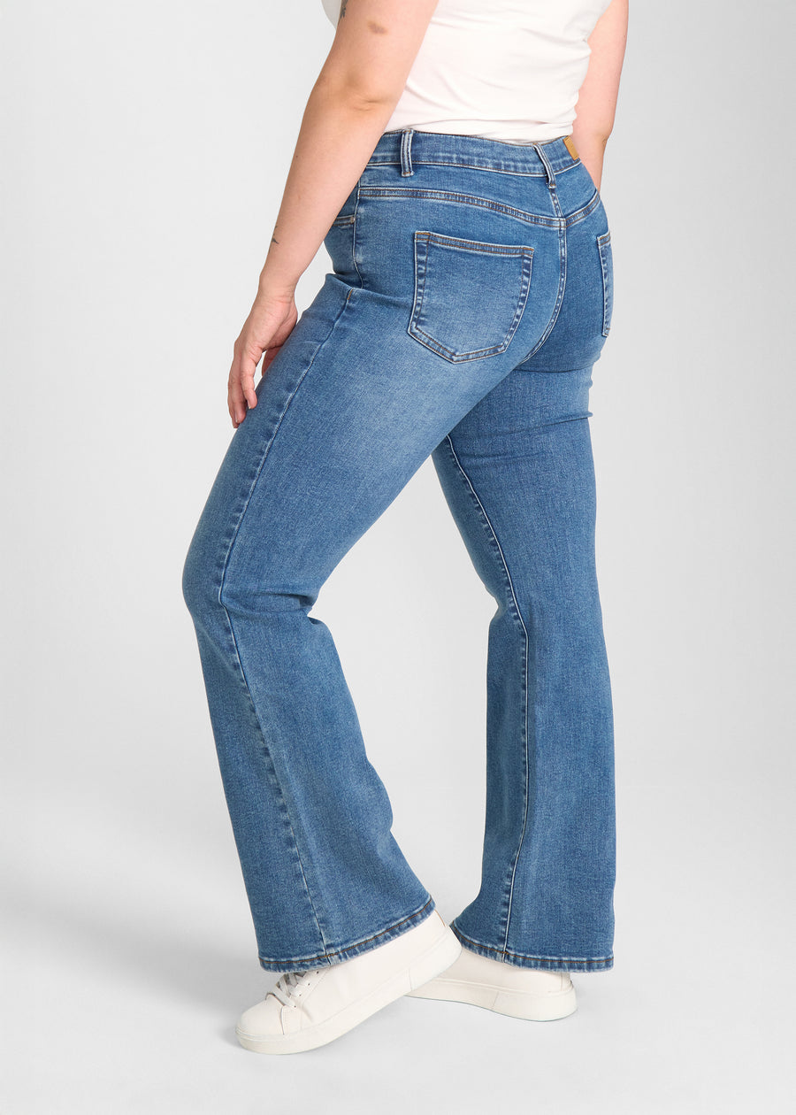 Blå denim bootcut jeans, modelnavn Rivers, som sidder tæt om hofter og lår med et let svaj fra knæet og ned, set fra siden på model på en plus-size model.