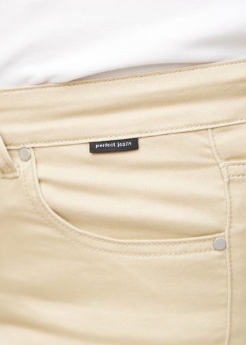 Close-up of beige jeans material with the Perfect Jeans logo label at the edge of the front pocket on a plus-size model.