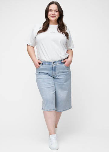 Full image of a plus-size model wearing loose, light blue denim capris in the colour Waves, reaching just below the knee.