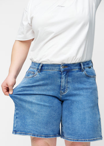 Plus-size model strækker de løse, klassisk blå denim shorts i farven Rivers ved hoften for at vise materialets stretch.