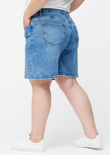 Løse, klassisk blå denim shorts i farven Rivers set fra siden på plus-size model, hvor den brede pasform og længden til lige over knæet er synlig.