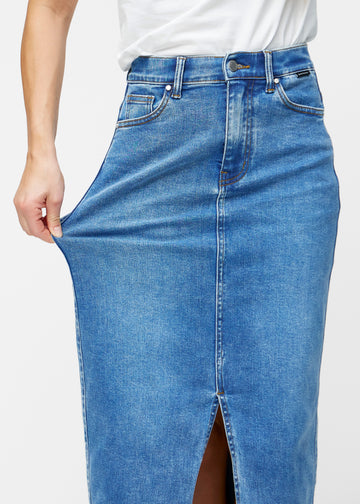 Model strækker den lange, klassisk blå denimnederdel i farven Rivers ved hoften for at vise materialets stretch.