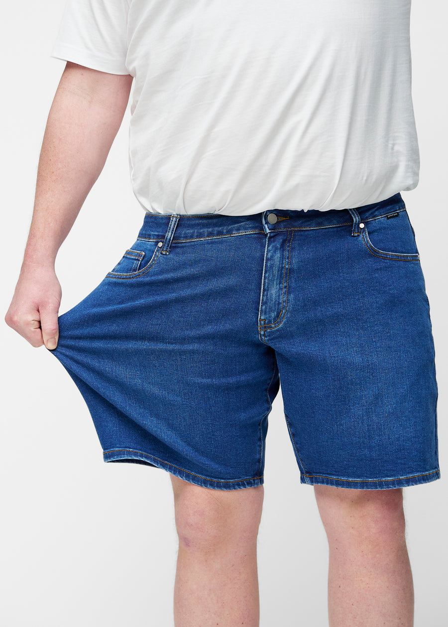 Plus-size model stretches the dark blue denim shorts for men in the colour Oceans at the thigh to show the stretch of the material.