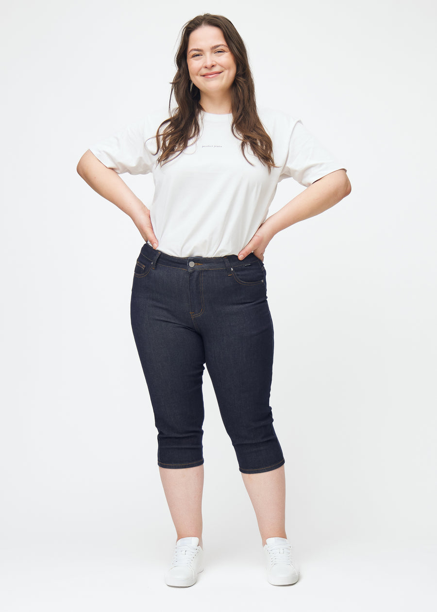 Full image of a plus-size model wearing deep dark blue denim capris for women in the colour Midnights with a skinny fit.