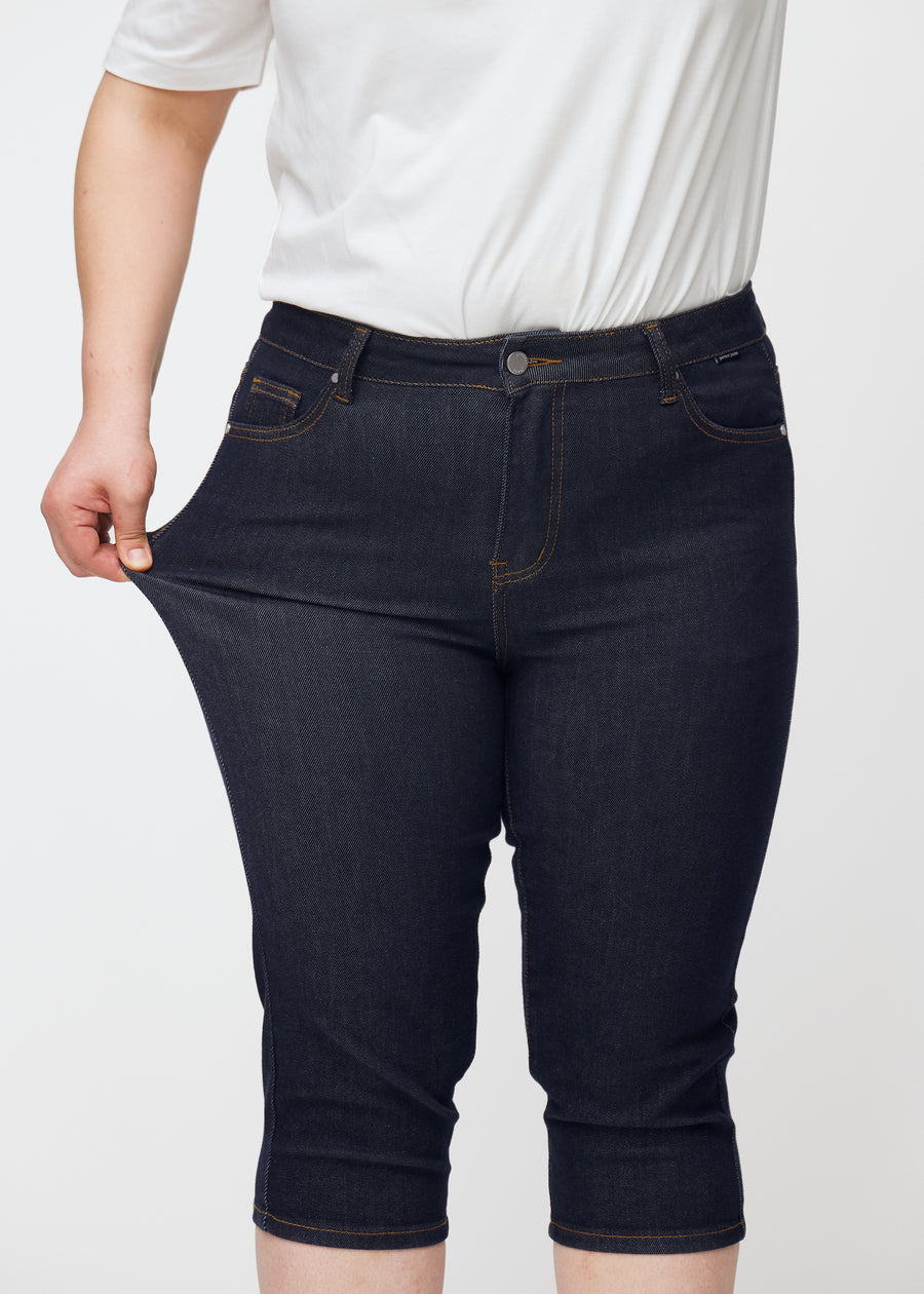 Plus-size model stretches deep dark blue denim capris for women in the colour Midnights at the thigh to show stretch.
