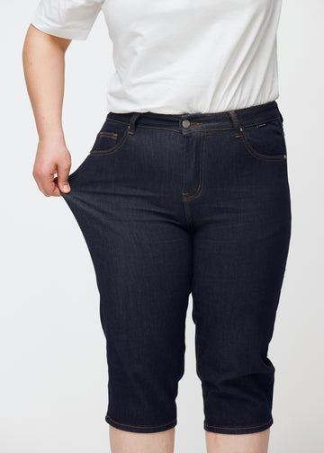 Plus-size model stretches deep dark blue denim capris for women in the colour Midnights at the thigh to show stretch.