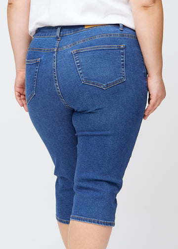 Slim capris for women in dark blue denim, color Oceans. Close-up from behind on a plus-size model, highlighting the back pockets and stitching.