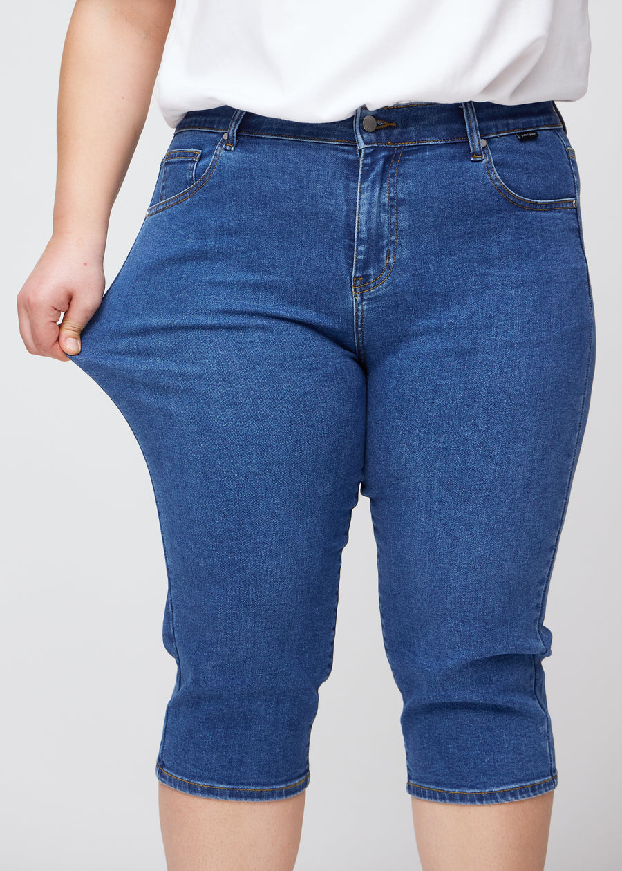 Slim capris for women in dark blue denim, color Oceans. A plus-size model pulls the fabric at the thigh to demonstrate stretch and flexibility.