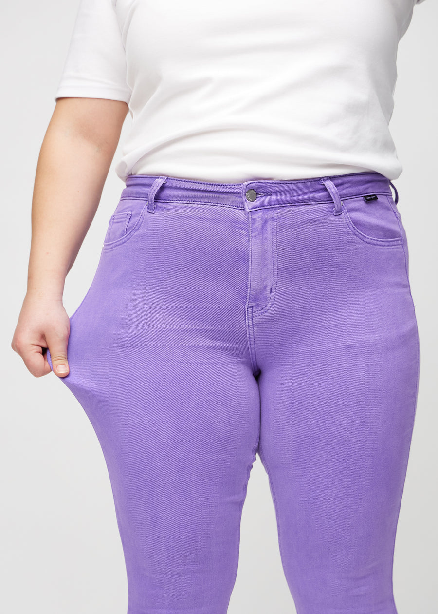 Skinny jeans for women in purple, color Lavenders. A plus-size model stretches the jeans at the thigh to show the fabric&