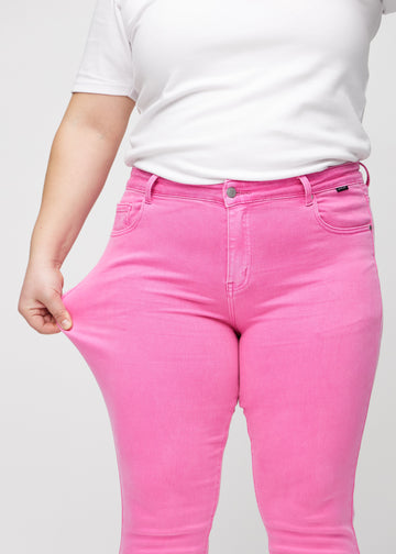 Slim jeans for women in light pink, color Flamingos. A plus-size model stretches the jeans at the thigh to show the fabric&