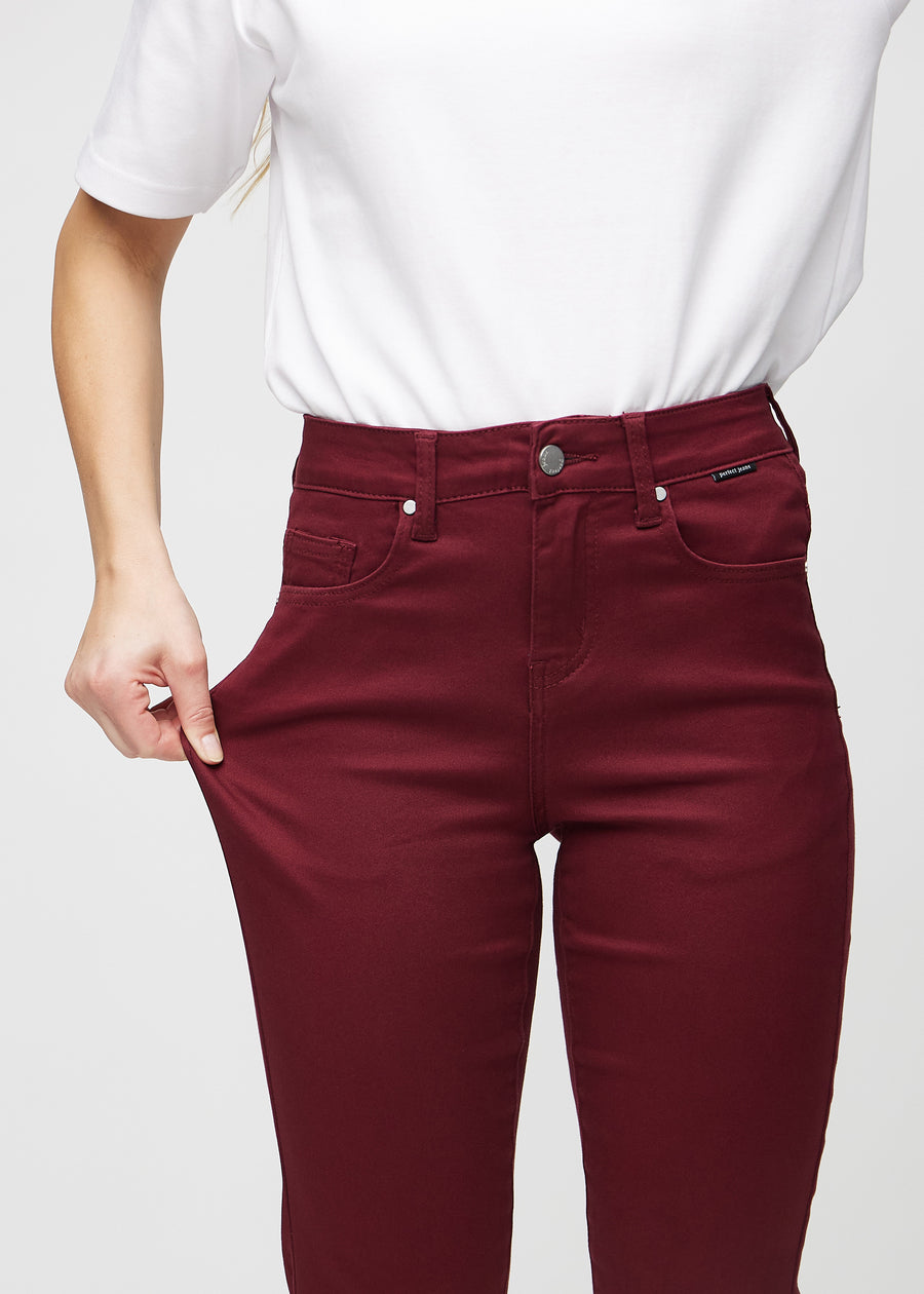 Slim jeans for women in dark red, color Santas. A model stretches the jeans at the thigh to show the fabric&