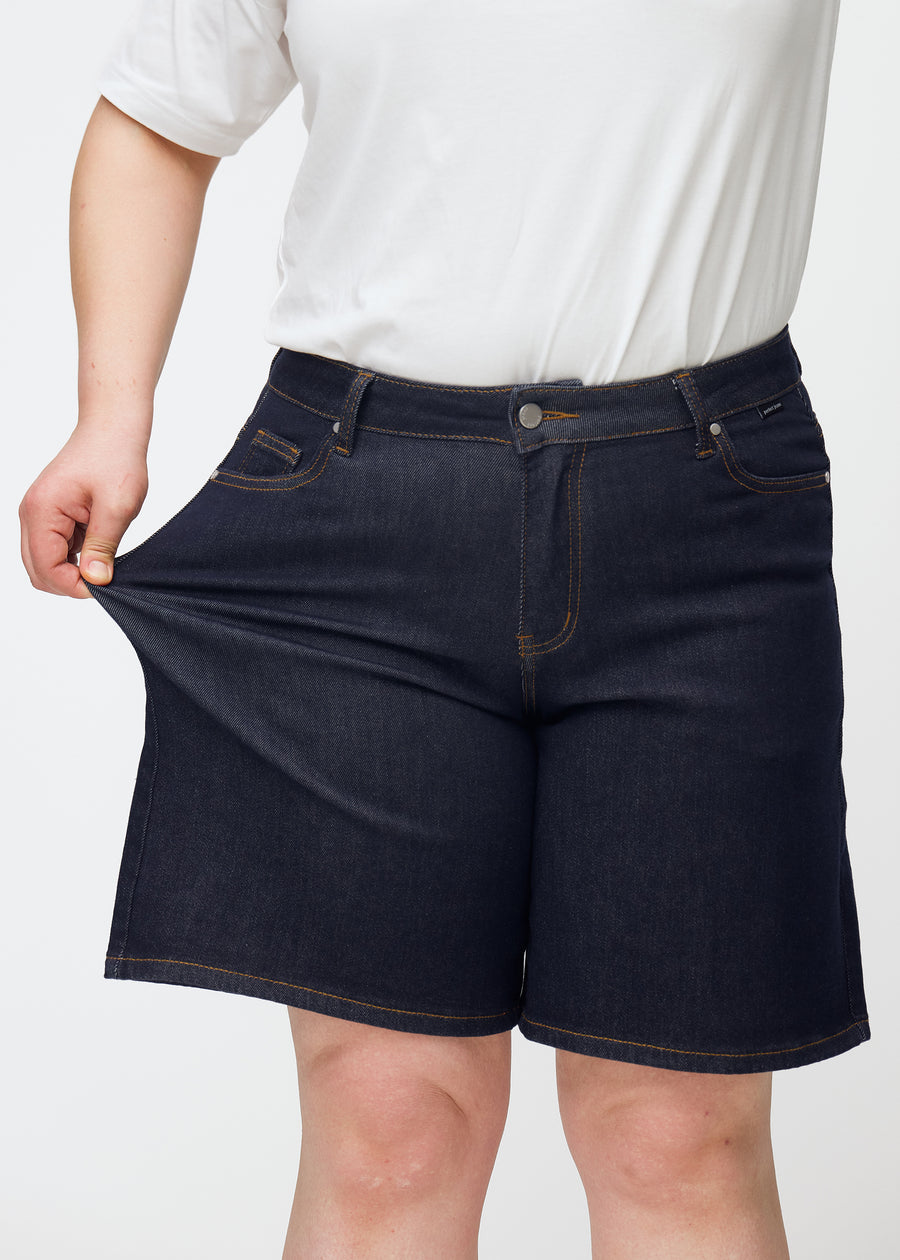 Plus-size model stretches deep dark blue denim shorts for women in the colour Midnights at the thigh to show stretch.